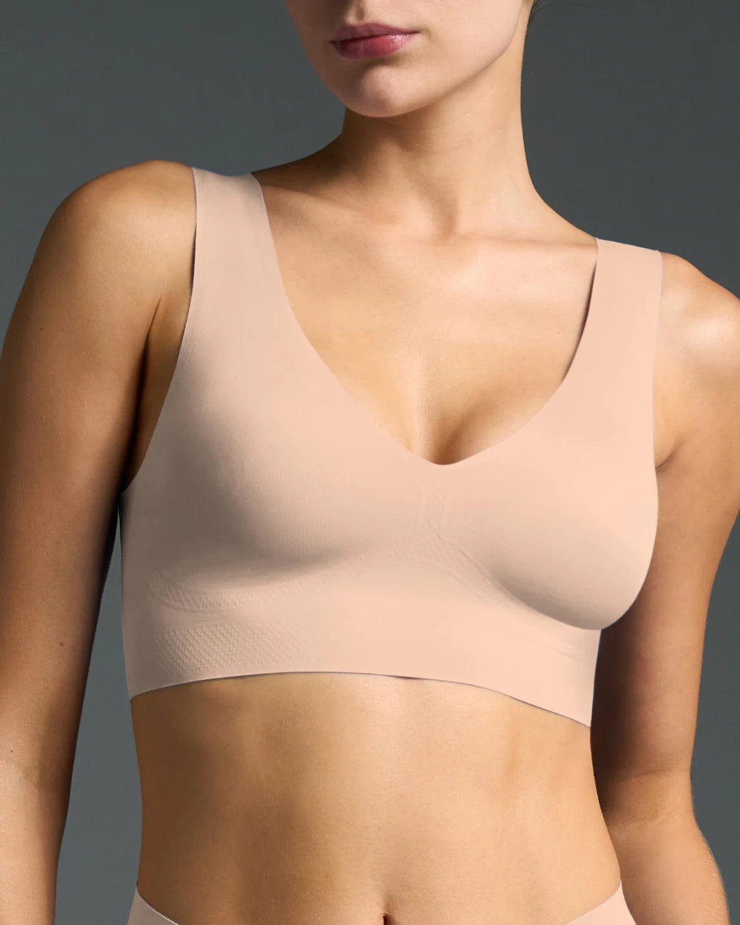 Model wearing nude seamless wireless bralette with wide straps and V-neckline against grey backdrop.